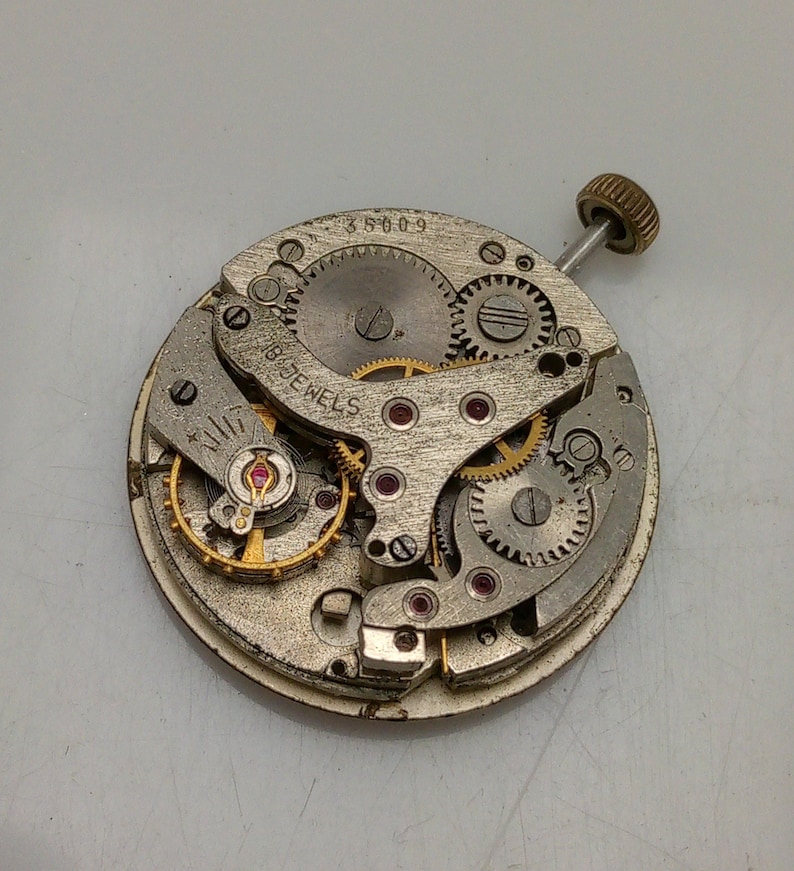 Op de afbeelding: Een zilveren uurwerk met een gouden opwindstift en de tekst "18 Jewels" en "35009".