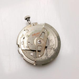 May include: A disassembled Seiko watch movement, showing intricate gears, screws, and a winding stem. The metal components display signs of wear. The text "SEIKO 21 JEWELS JAPAN" is visible on the movement.