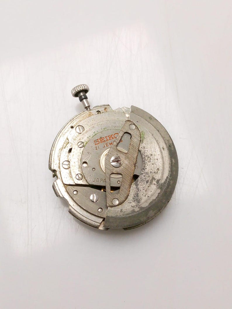 May include: A disassembled Seiko watch movement, showing intricate gears, screws, and a winding stem. The metal components display signs of wear. The text "SEIKO 21 JEWELS JAPAN" is visible on the movement.