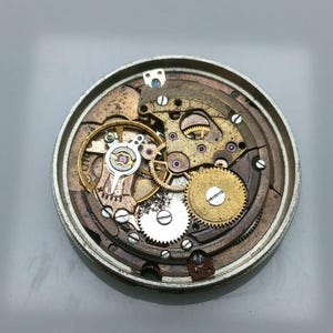 May include: Close-up of a disassembled watch mechanism. The intricate internal components, including gears, springs, and screws, are visible. The metal parts are in shades of gold, silver, and bronze, showcasing the watch's inner workings.