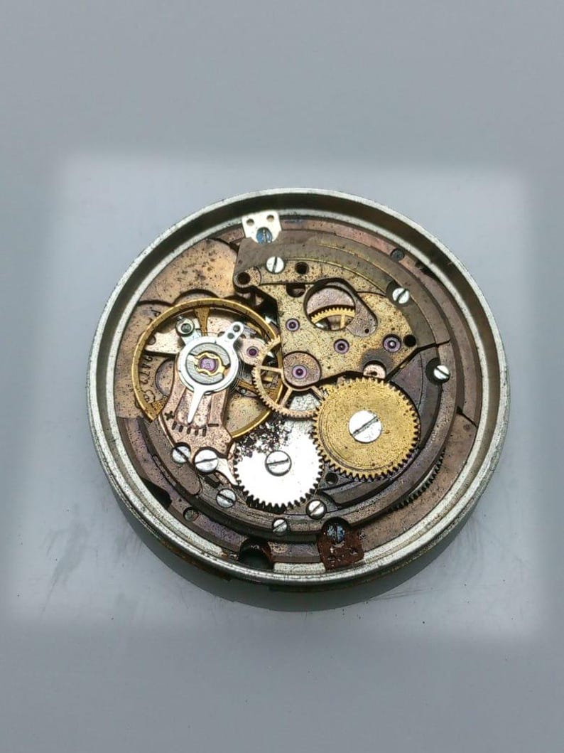 May include: Close-up of a disassembled watch mechanism. The intricate internal components, including gears, springs, and screws, are visible. The metal parts are in shades of gold, silver, and bronze, showcasing the watch's inner workings.