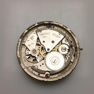 May include: Close-up of a vintage Orient watch movement. The silver-toned mechanism features intricate gears, jewels, and the text "ORIENT 17J. 20321". The watch components are housed within a circular frame.