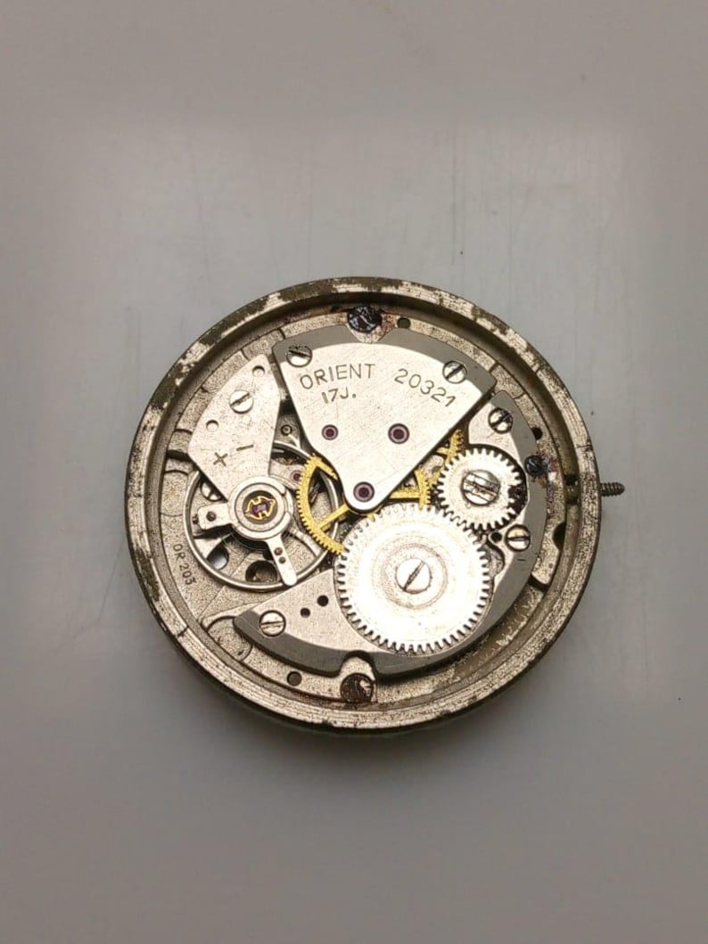May include: Close-up of a vintage Orient watch movement. The silver-toned mechanism features intricate gears, jewels, and the text "ORIENT 17J. 20321". The watch components are housed within a circular frame.