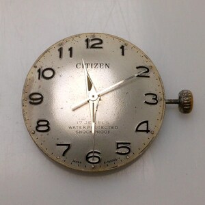 May include: A vintage Citizen watch face with a silver dial, black numerals, and silver hands. The watch face includes the text "CITIZEN", "17 JEWELS", "WATER PROTECTED", and "SHOCK PROOF". A winding stem is attached to the side.