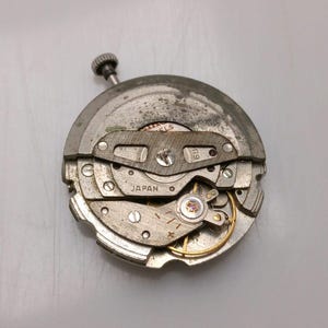 May include: A detailed view of a vintage watch movement, showcasing its complex internal mechanisms. The metal components are silver and grey. The word "JAPAN" is stamped on the movement. The crown is visible at the top.
