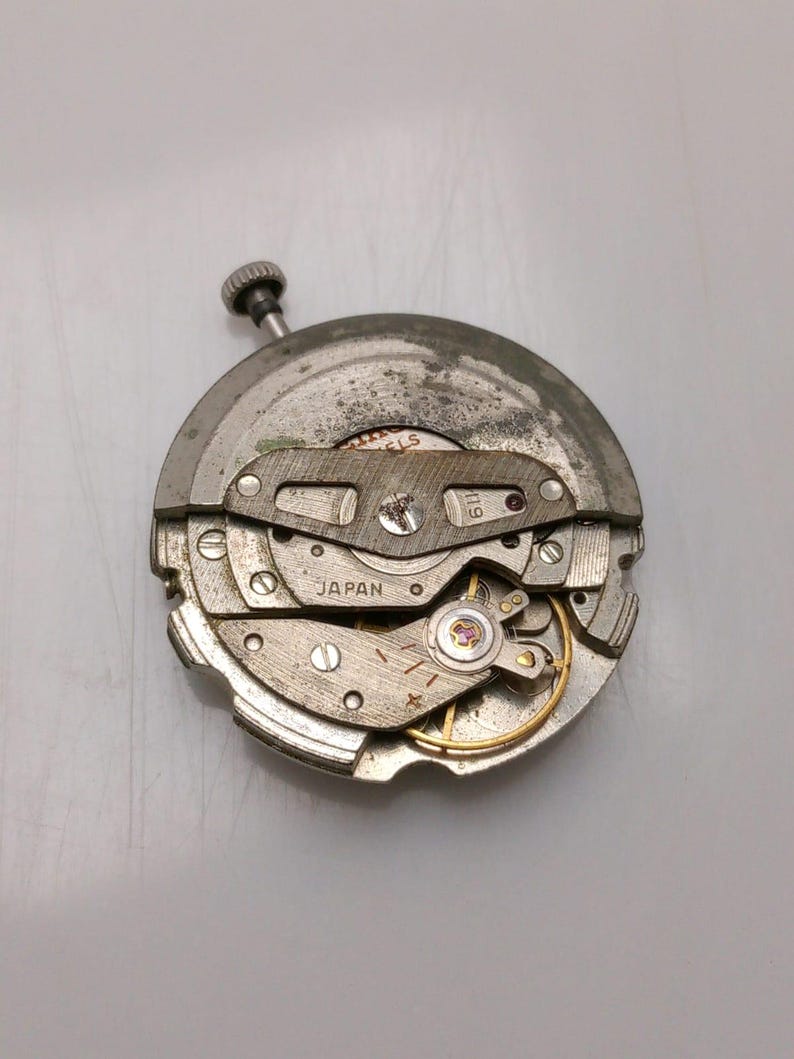 May include: A detailed view of a vintage watch movement, showcasing its complex internal mechanisms. The metal components are silver and grey. The word "JAPAN" is stamped on the movement. The crown is visible at the top.