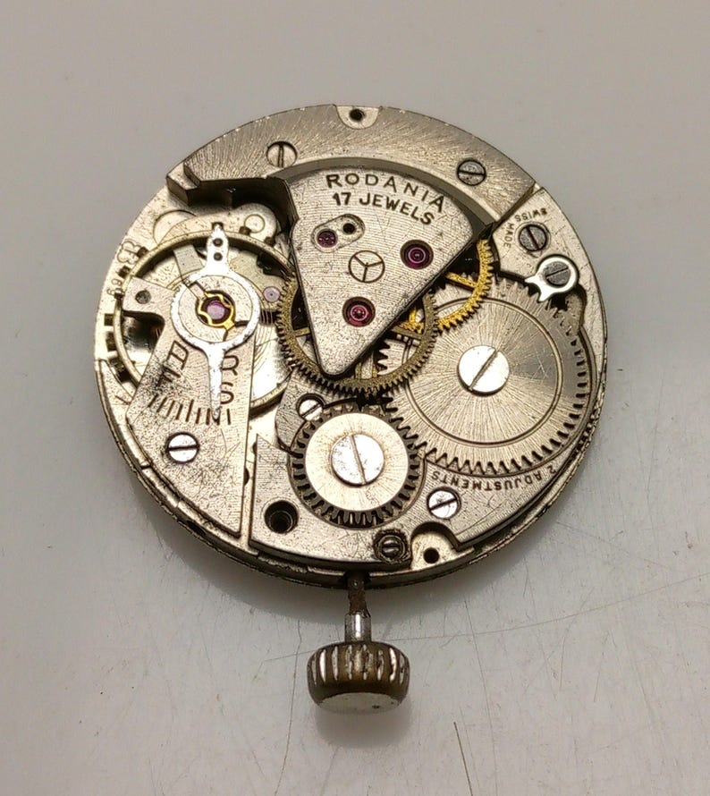 Op de afbeelding: Close-up van een zilveren uurwerk met de tekst "Rodania 17 Jewels" op de wijzerplaat. Het uurwerk is gedemonteerd en toont de tandwielen en andere interne componenten.