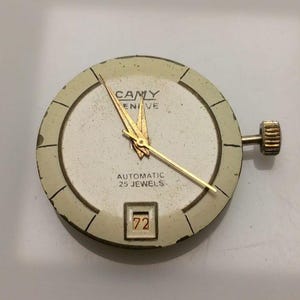 May include: A vintage watch face with a cream-colored dial and gold hands. The watch face has the text "CAMY GENEVE AUTOMATIC 25 JEWELS" and a date window displaying the number "72". The watch has a gold crown.