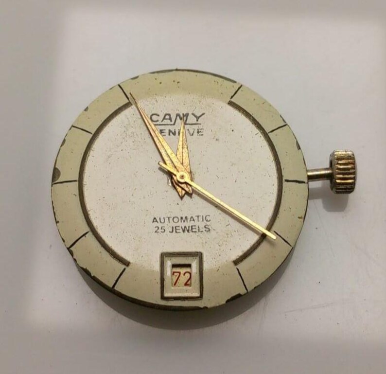 May include: A vintage watch face with a cream-colored dial and gold hands. The watch face has the text "CAMY GENEVE AUTOMATIC 25 JEWELS" and a date window displaying the number "72". The watch has a gold crown.
