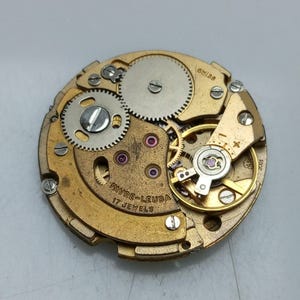 Puede incluir: Un movimiento de reloj de color dorado con el texto "Favre-Leuba 17 Jewels" y "Swiss" impreso en él.