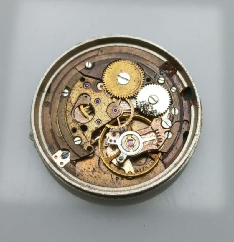 May include: Close-up of a vintage watch movement, showcasing intricate gears, springs, and screws. The mechanism features brass and silver-toned components, with a circular design. The watch parts are arranged within a metal casing, revealing the inner workings of the timepiece.