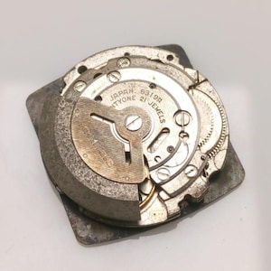 Puede incluir: Primer plano de un movimiento de reloj vintage con engranajes y componentes intrincados. El mecanismo del reloj est&aacute; marcado como "Japan Twenty-one Jewels" y "6319A". Las piezas met&aacute;licas son de color plateado y gris.
