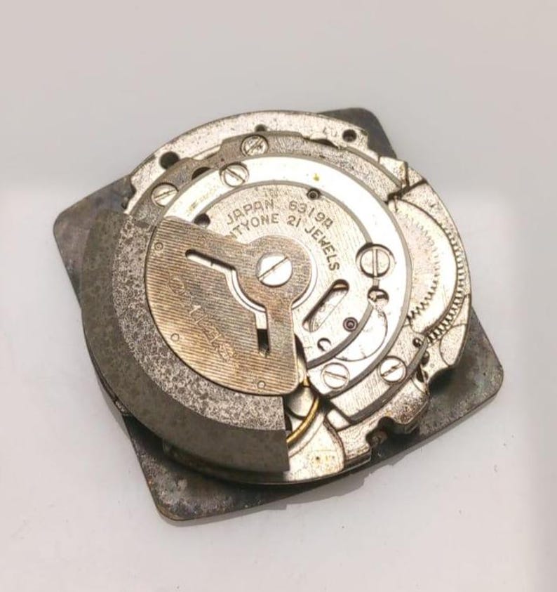 Puede incluir: Primer plano de un movimiento de reloj vintage con engranajes y componentes intrincados. El mecanismo del reloj est&aacute; marcado como "Japan Twenty-one Jewels" y "6319A". Las piezas met&aacute;licas son de color plateado y gris.