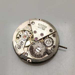 May include: A close-up of a disassembled Roamer watch movement. The silver-toned mechanism features intricate gears, jewels, and the text "ROAMER 17 JEWELS SWISS MADE". The watch movement is circular and shows signs of wear.