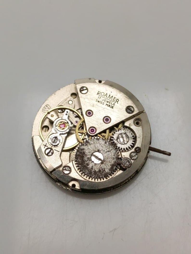 May include: A close-up of a disassembled Roamer watch movement. The silver-toned mechanism features intricate gears, jewels, and the text "ROAMER 17 JEWELS SWISS MADE". The watch movement is circular and shows signs of wear.