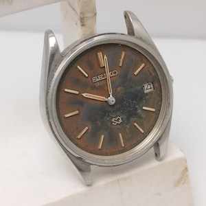 Könnte beinhalten: Vintage Seiko-Uhr mit silberfarbenem Gehäuse und einem verwitterten braunen Zifferblatt. Das Zifferblatt hat goldfarbene Stundenmarkierungen und Zeiger, mit einem Datumsfenster bei 3 Uhr. Die Uhr hat kein Armband.