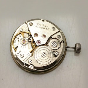 May include: A close-up of a Citizen watch movement with the text "CITIZEN 21 JEWELS". The watch mechanism features various gears, screws, and a winding stem. The movement is silver and gold in color.