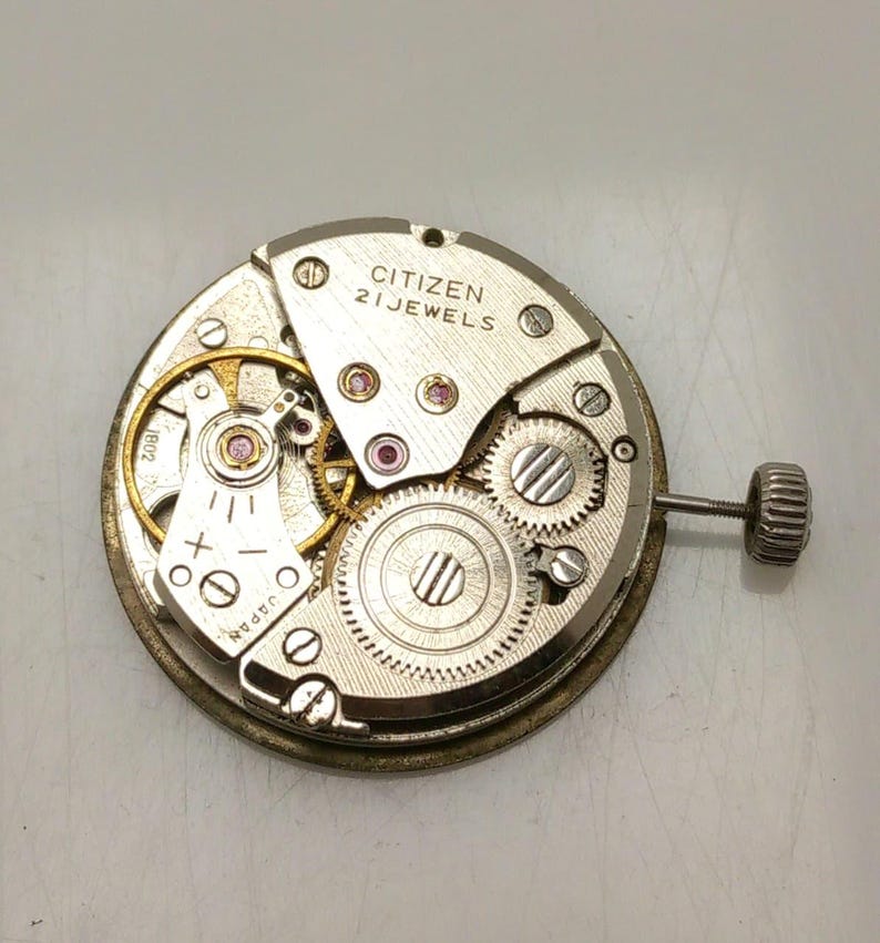 May include: A close-up of a Citizen watch movement with the text "CITIZEN 21 JEWELS". The watch mechanism features various gears, screws, and a winding stem. The movement is silver and gold in color.