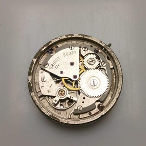 May include: Close-up of an Orient watch movement, showcasing intricate gears, springs, and jewels. The watch mechanism is housed in a circular, aged metal casing. The text "ORIENT 17J. 20321" is visible on the watch face.