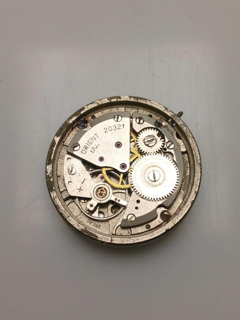 May include: Close-up of an Orient watch movement, showcasing intricate gears, springs, and jewels. The watch mechanism is housed in a circular, aged metal casing. The text "ORIENT 17J. 20321" is visible on the watch face.
