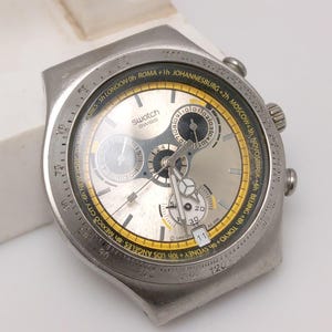 May include: A silver-toned Swatch Swiss watch with a round face. The watch features a silver dial with black and yellow accents, including a world time bezel. The watch has multiple sub-dials and a date display.