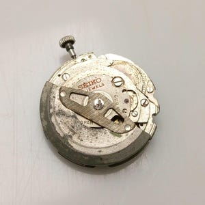 May include: A close-up of a Seiko watch movement. The silver mechanism features the brand name and the word "JEWELS" printed on the face. A small, textured crown is visible at the top, and various gears and components are arranged throughout.