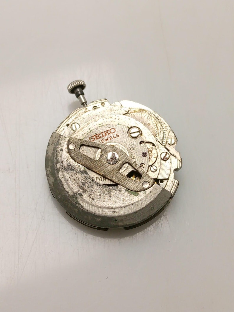 May include: A close-up of a Seiko watch movement. The silver mechanism features the brand name and the word "JEWELS" printed on the face. A small, textured crown is visible at the top, and various gears and components are arranged throughout.