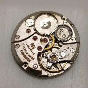 May include: Close-up of a vintage Longines watch movement. The silver-toned mechanism features intricate gears, ruby jewels, and the text "17 Jewels Swiss". The watch movement is a complex arrangement of metal components.