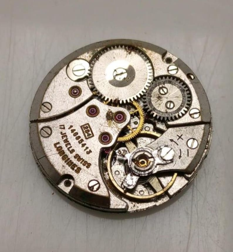 May include: Close-up of a vintage Longines watch movement. The silver-toned mechanism features intricate gears, ruby jewels, and the text "17 Jewels Swiss". The watch movement is a complex arrangement of metal components.