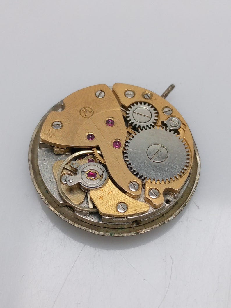 Puede incluir: Primer plano de un movimiento de reloj dorado con engranajes visibles y un peque&ntilde;o tornillo. El movimiento est&aacute; sobre un fondo blanco.