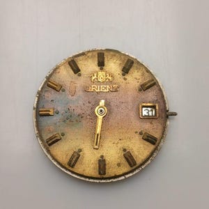 May include: A vintage Orient watch face with a gold-toned dial and hands. The dial features rectangular hour markers, the Orient logo, and a date window displaying the number 31. The watch face has a weathered appearance, suggesting age and use.