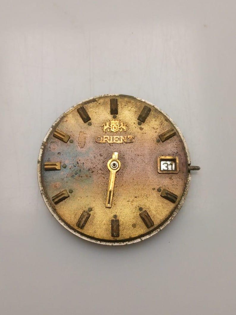 May include: A vintage Orient watch face with a gold-toned dial and hands. The dial features rectangular hour markers, the Orient logo, and a date window displaying the number 31. The watch face has a weathered appearance, suggesting age and use.