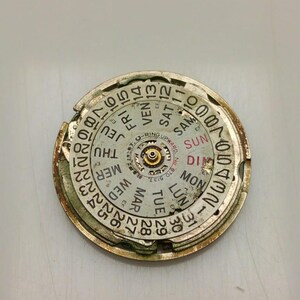 May include: A circular watch component with a gold-colored frame and a white face. The face displays the days of the week and numbers for the date. The component has a central mechanism and is slightly damaged.