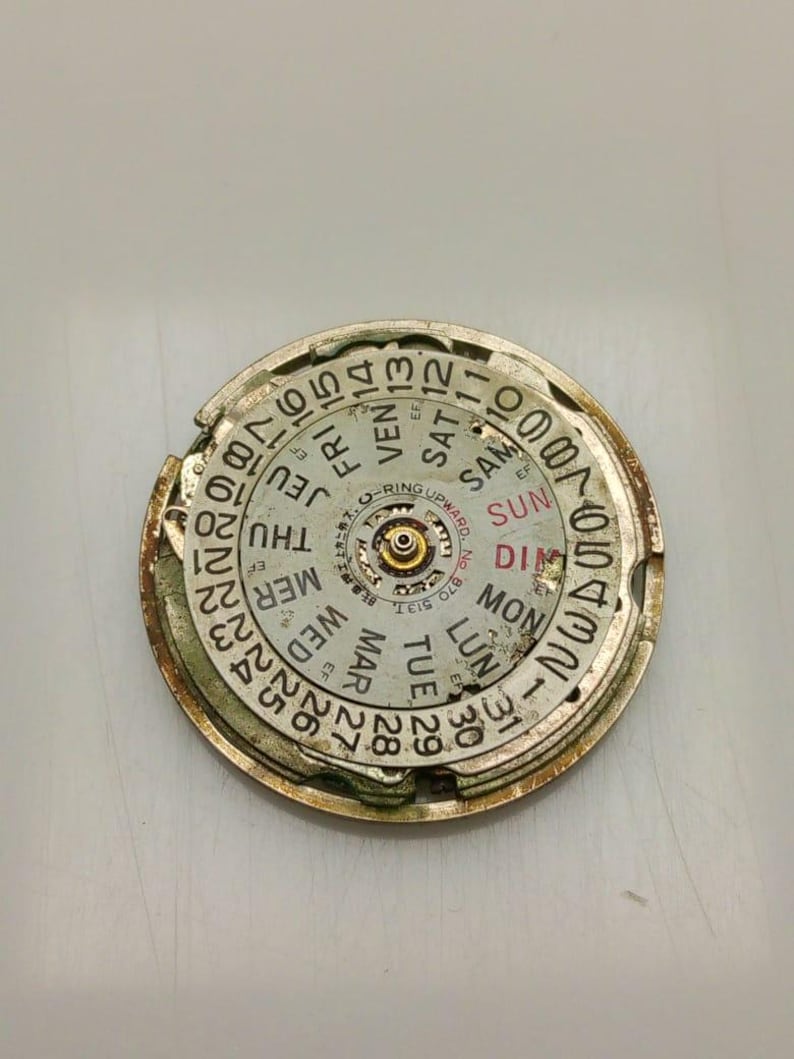 May include: A circular watch component with a gold-colored frame and a white face. The face displays the days of the week and numbers for the date. The component has a central mechanism and is slightly damaged.