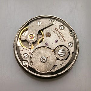 May include: A close-up of a vintage Seiko watch movement. The mechanism features silver gears, screws, and plates, with gold accents. The text "SEIKOSHA 17 JEWELS" is visible. The watch movement is a circular shape.