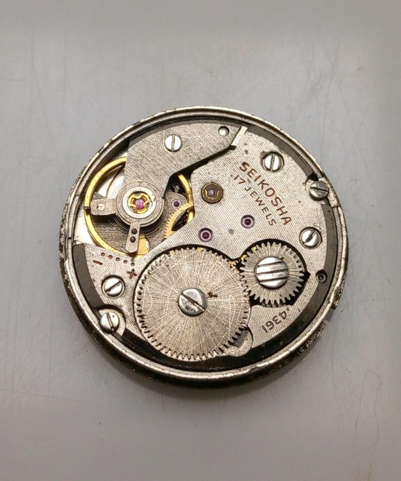 May include: A close-up of a vintage Seiko watch movement. The mechanism features silver gears, screws, and plates, with gold accents. The text "SEIKOSHA 17 JEWELS" is visible. The watch movement is a circular shape.
