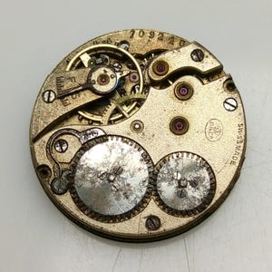 Pode incluir: Close-up de um movimento de rel&oacute;gio prateado com engrenagens intrincadas e uma inscri&ccedil;&atilde;o 'SWISS MADE'. O movimento tem um design circular com uma abertura central e v&aacute;rias engrenagens menores ao redor.