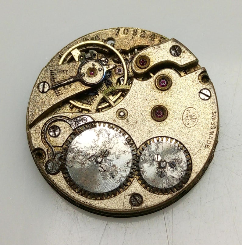 Pode incluir: Close-up de um movimento de rel&oacute;gio prateado com engrenagens intrincadas e uma inscri&ccedil;&atilde;o 'SWISS MADE'. O movimento tem um design circular com uma abertura central e v&aacute;rias engrenagens menores ao redor.