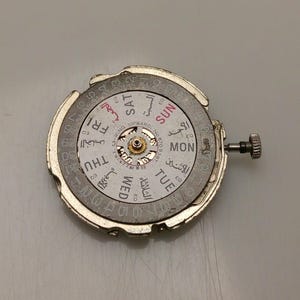 May include: A silver watch movement with a white dial showing the days of the week in English and Arabic. The dial also has the numbers 1-31. A small crown is visible on the right side.