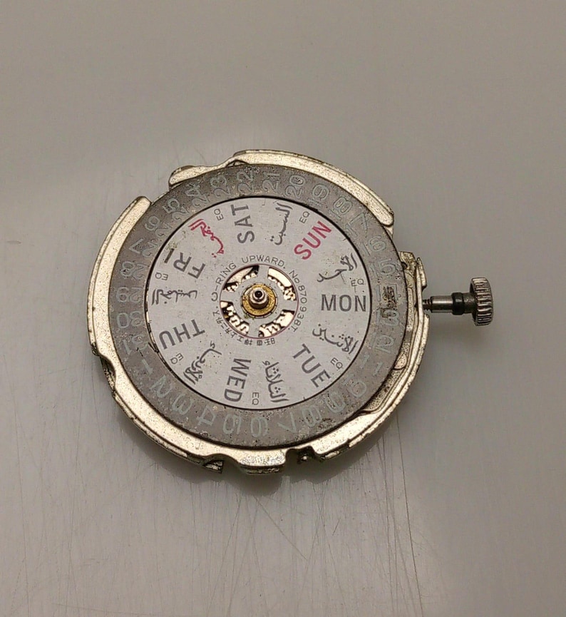 May include: A silver watch movement with a white dial showing the days of the week in English and Arabic. The dial also has the numbers 1-31. A small crown is visible on the right side.