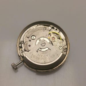 Pode incluir: Um close-up de um movimento de rel&oacute;gio Orient 21J 46943 Japan prateado. O mecanismo do rel&oacute;gio &eacute; vis&iacute;vel, incluindo engrenagens, parafusos e uma coroa. O movimento do rel&oacute;gio &eacute; circular e tem uma estrutura interna complexa.