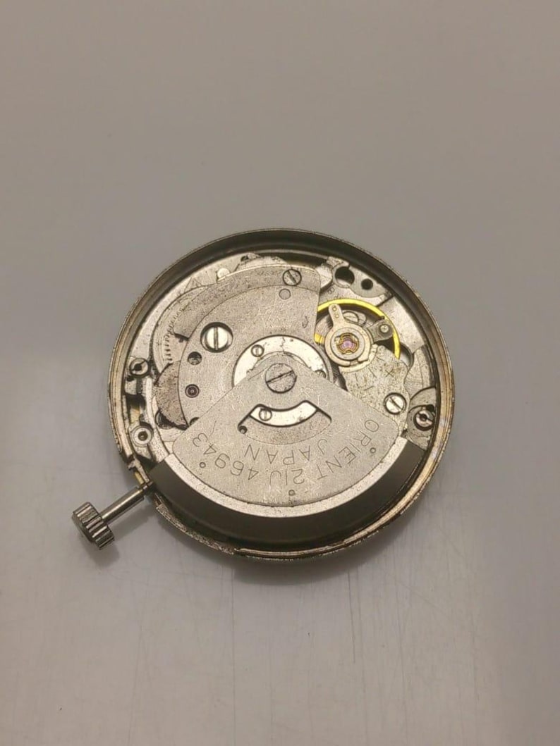 Pode incluir: Um close-up de um movimento de rel&oacute;gio Orient 21J 46943 Japan prateado. O mecanismo do rel&oacute;gio &eacute; vis&iacute;vel, incluindo engrenagens, parafusos e uma coroa. O movimento do rel&oacute;gio &eacute; circular e tem uma estrutura interna complexa.