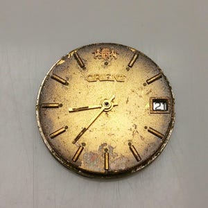 Puede incluir: Una esfera de reloj Orient vintage con acabado dorado. Presenta agujas doradas, marcadores de hora y el nombre de la marca "ORIENT". La fecha se muestra como "21". La esfera del reloj muestra signos de desgaste y envejecimiento.