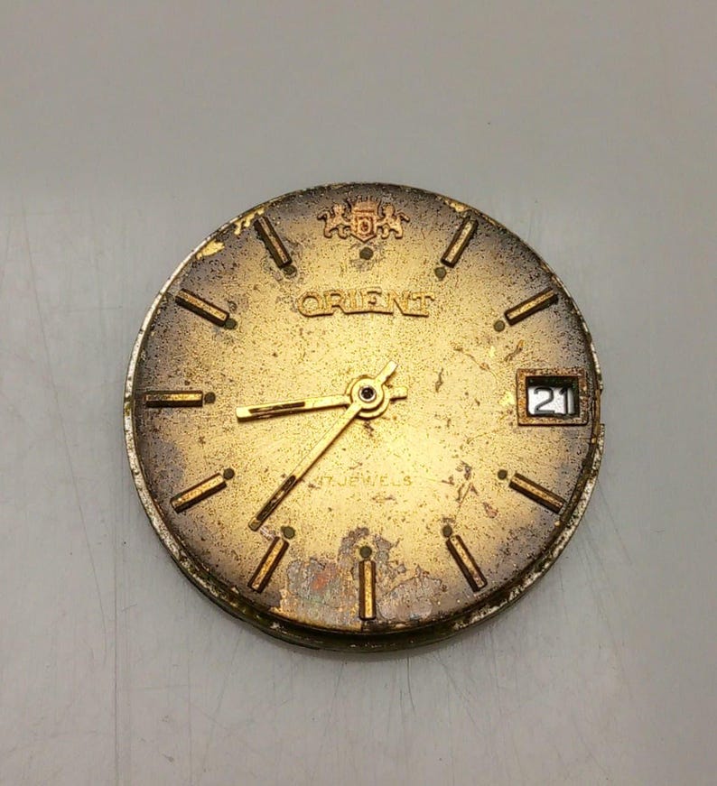 Puede incluir: Una esfera de reloj Orient vintage con acabado dorado. Presenta agujas doradas, marcadores de hora y el nombre de la marca "ORIENT". La fecha se muestra como "21". La esfera del reloj muestra signos de desgaste y envejecimiento.