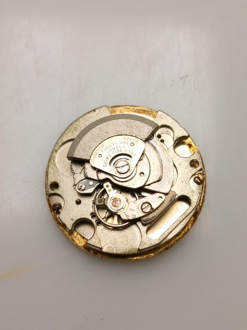 May include: Close-up of a watch movement with intricate gears and components. The movement has a gold-colored outer ring and silver-colored internal parts. The text "Twenty-one Jewels Seiko Time Corp" is visible.