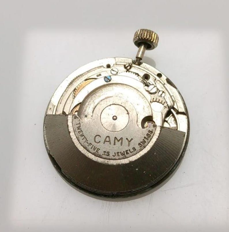 May include: A close-up of a vintage watch movement. The silver-toned mechanism features the brand name "CAMY" and the text "TWENTY-FIVE JEWELS SWISS". The watch movement has a gold-colored crown and intricate internal components.