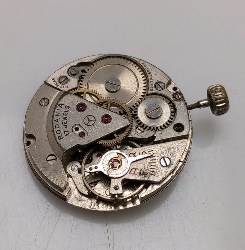 Op de afbeelding: Een zilverkleurig uurwerk met de tekst "Rodania 17 Jewels" en "2 Adjustments". Het uurwerk is gedemonteerd en toont de interne werking van het horloge.