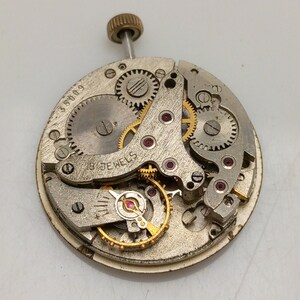 Op de afbeelding: Een close-up van een zilveren uurwerk met gouden tandwielen en een opwindas. Het uurwerk heeft 18 juwelen en de tekst "318009" is zichtbaar aan de zijkant.