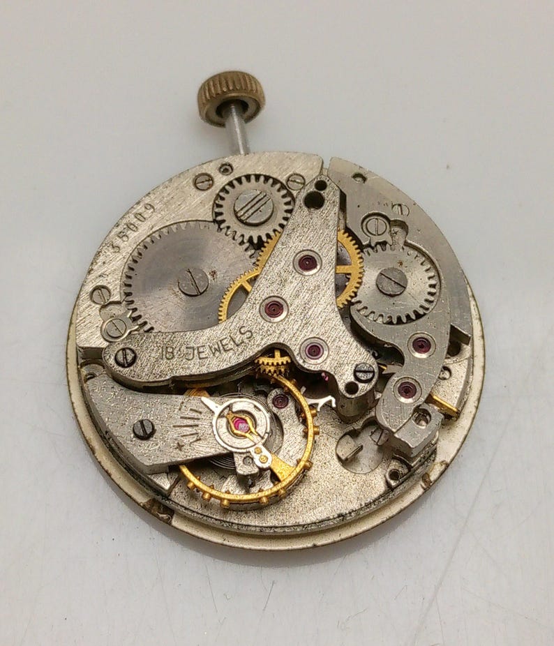 Op de afbeelding: Een close-up van een zilveren uurwerk met gouden tandwielen en een opwindas. Het uurwerk heeft 18 juwelen en de tekst "318009" is zichtbaar aan de zijkant.