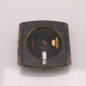 Puede incluir: Una esfera de reloj Seiko vintage con esfera negra y detalles dorados. La esfera cuadrada tiene una esfera circular en el centro. La palabra "AUTOMATIC" est&aacute; impresa en la esfera. El logotipo de Seiko es dorado. Es una pieza de repuesto.